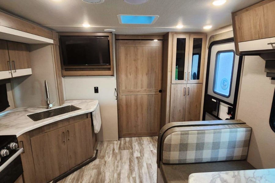 Modern RV rental interior: kitchen with sink & stove, dinette with plaid cushions, TV, and wood-look flooring.
