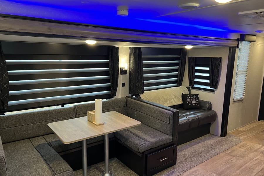 Modern rv rental interior: A cozy dinette with booth seating, blue accent lights, privacy blinds, and sleek wood-look flooring.