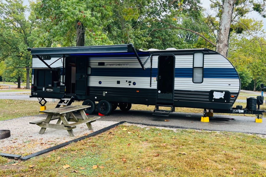 An RV rental is parked at a treed campsite on a gravel pad, with its awning out, alongside a picnic table and fire pit.