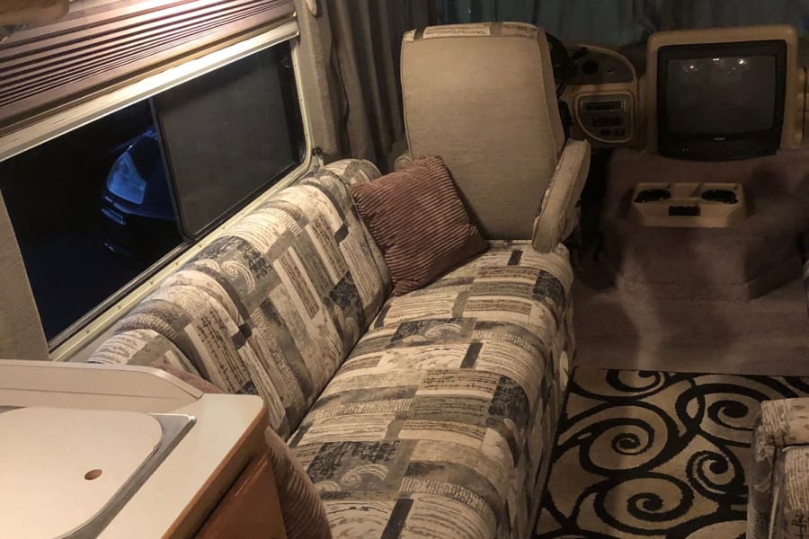 Cozy RV rental interior, showcasing a patterned sofa, compact kitchenette, and driver's cabin with an old-style TV.