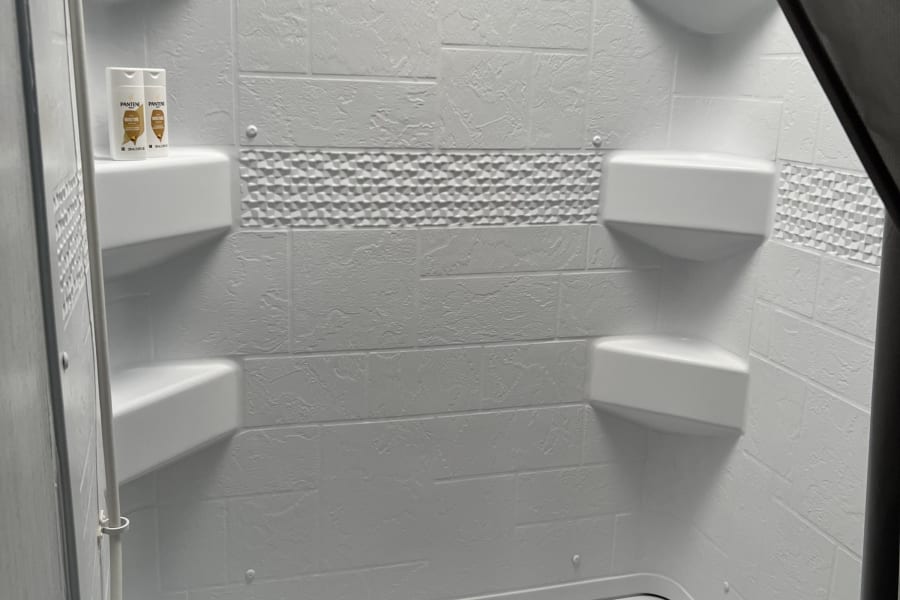 Inside a compact RV rental: a clean, white shower and tub with textured walls, built-in shelves, and bathing essentials.
