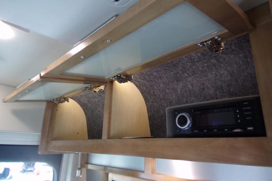 RV rental overhead cabinets with an integrated stereo system. Features light wood, opaque glass doors, & felt-lined storage.
