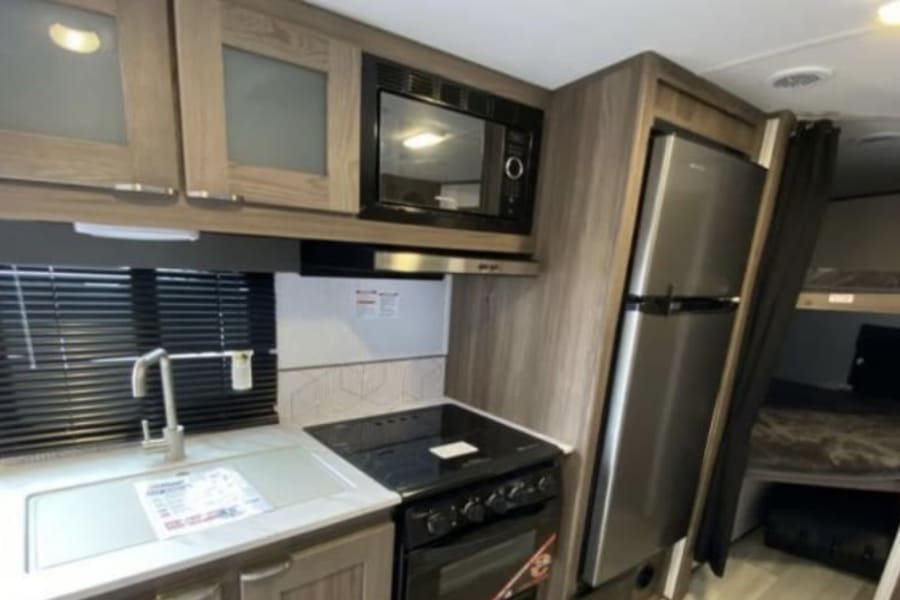 Modern RV rental kitchen with microwave, stove, sink, and fridge. Adjacent area includes bunk beds. Explore comfortable travel!