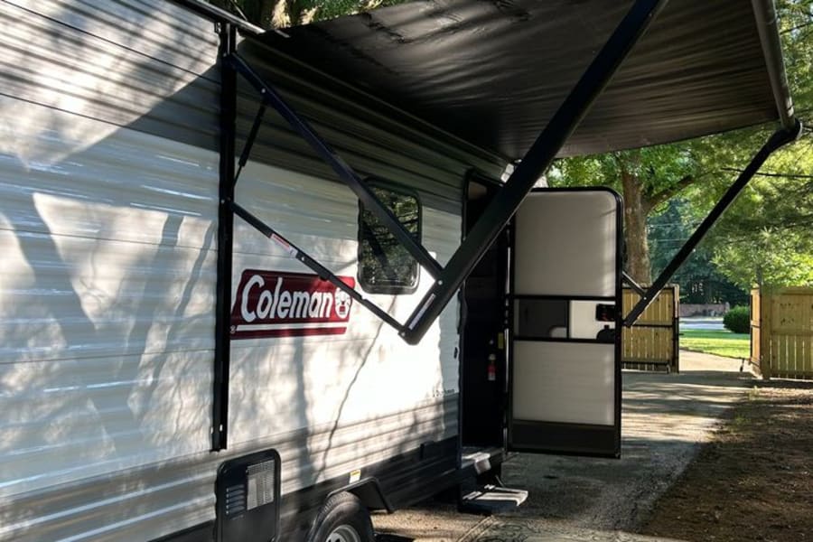 Coleman RV rental with an extended awning and large outdoor rug, set up at a sunny campsite.