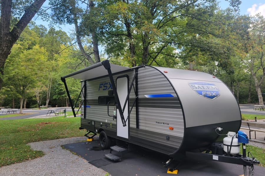 A grey Salem FSX RV rental travel trailer with awning extended, parked at a serene, treed campground.