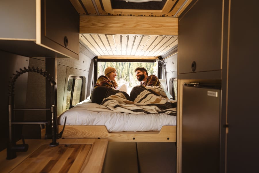 Cozy RV rental: A couple relaxes on a comfortable bed with a forest view. The van's interior features a kitchen sink and wood accents.