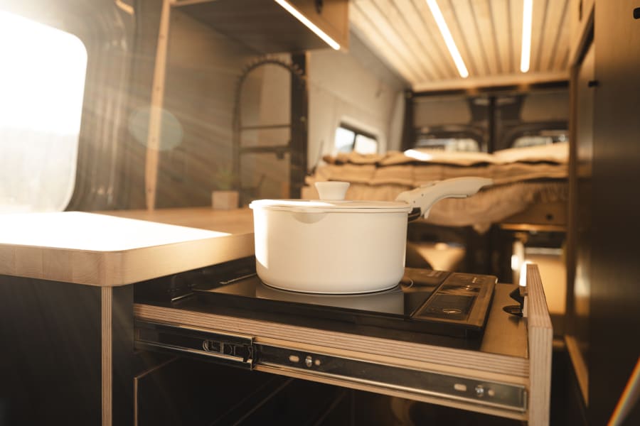 Modern RV rental interior: a white pot on a pull-out induction cooktop in a sunlit kitchen, with a cozy bed in the back.