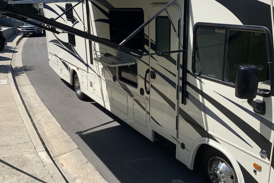 Cream RV rental, with black stripes & extended awning, parked on a sunny street, showing its side features.