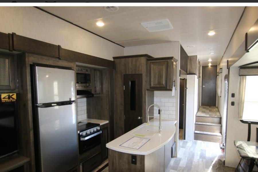 Spacious RV rental interior with a modern kitchen, featuring a tall fridge, stove, and island sink. Light walls and wood accents.