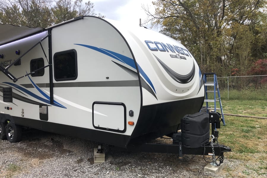 White and blue RV rental parked on gravel with an extended awning and LED lights, ready for an outdoor adventure.