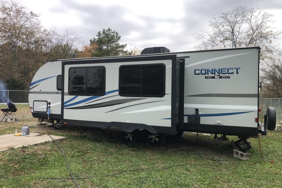 A grey 'Connect' travel trailer RV rental, with slide-out extended, is parked on grass with a grill, ready for camping.