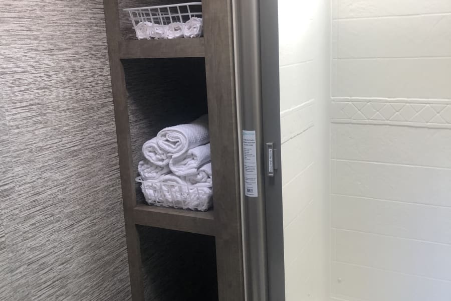 RV rental bathroom features a shower, built-in shelves with fresh towels, and wall hooks. Part of a toilet is visible.