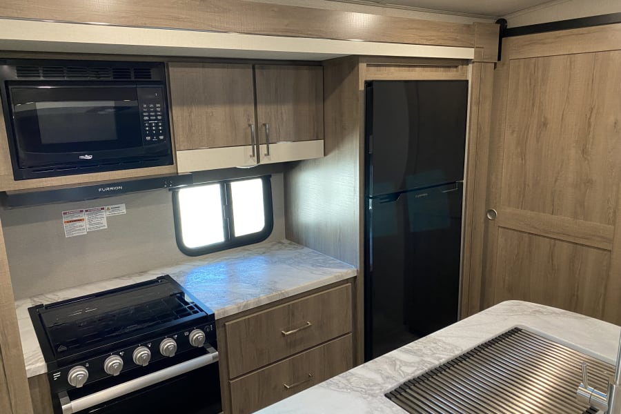 Modern RV rental kitchen with microwave, Furrion stove, refrigerator, sink, and light wood cabinetry.