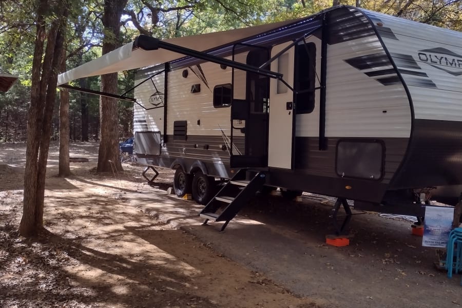 An RV rental is set up at a shaded campsite, featuring an extended awning & steps in a wooded area.