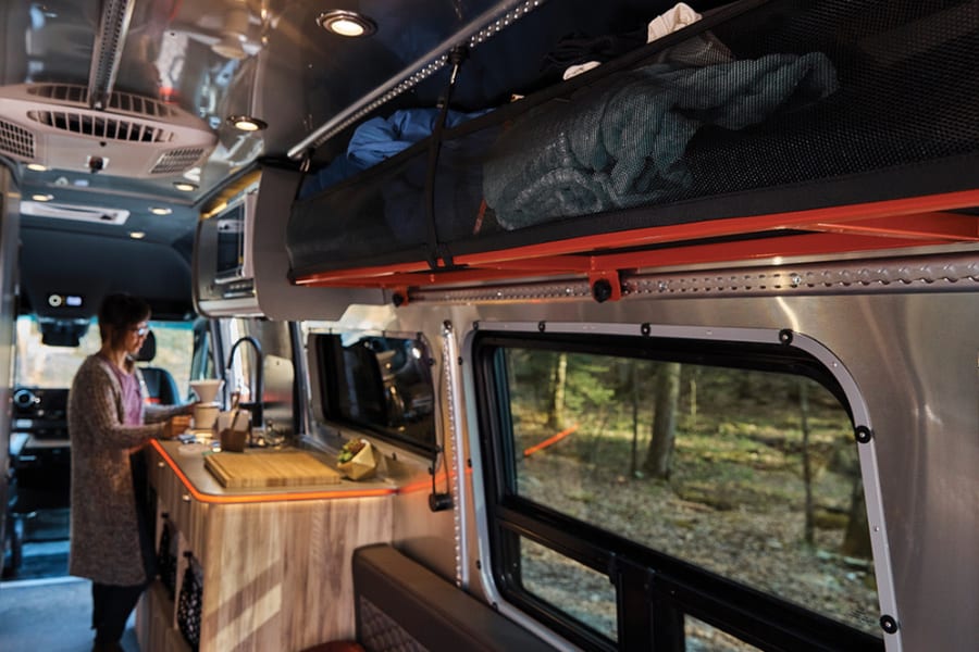 Experience an RV rental: a cozy interior with a person making coffee, forest views, and smart storage.