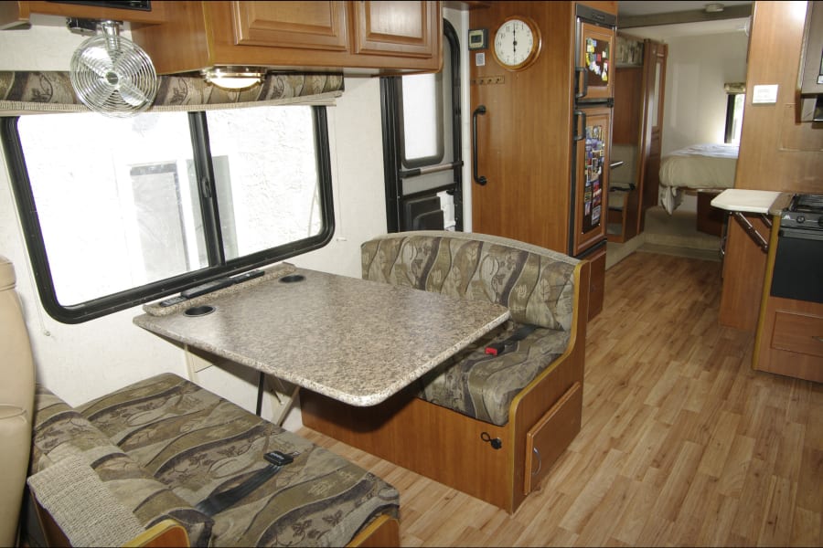 Inside an rv rental: Dinette with patterned benches, window, kitchen, and bedroom visible. Wood-look flooring.