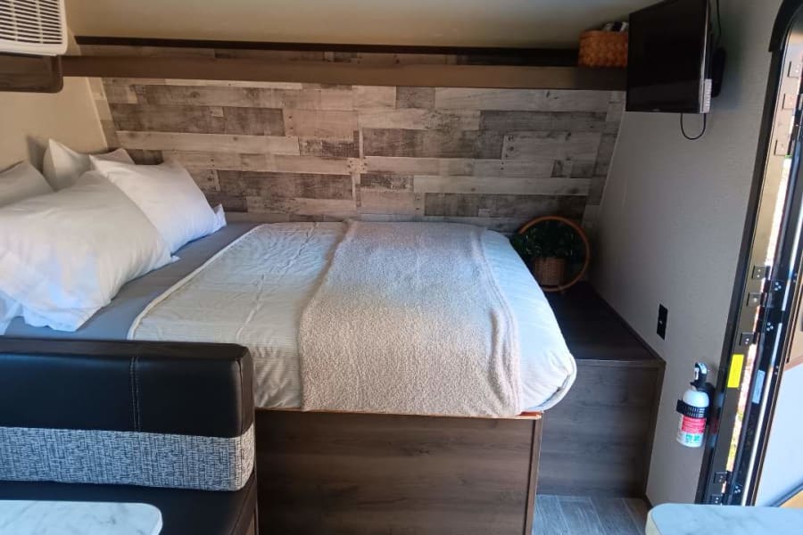 Explore this cozy RV rental interior: a comfortable bed, stylish wood-paneled headboard, and compact dinette.
