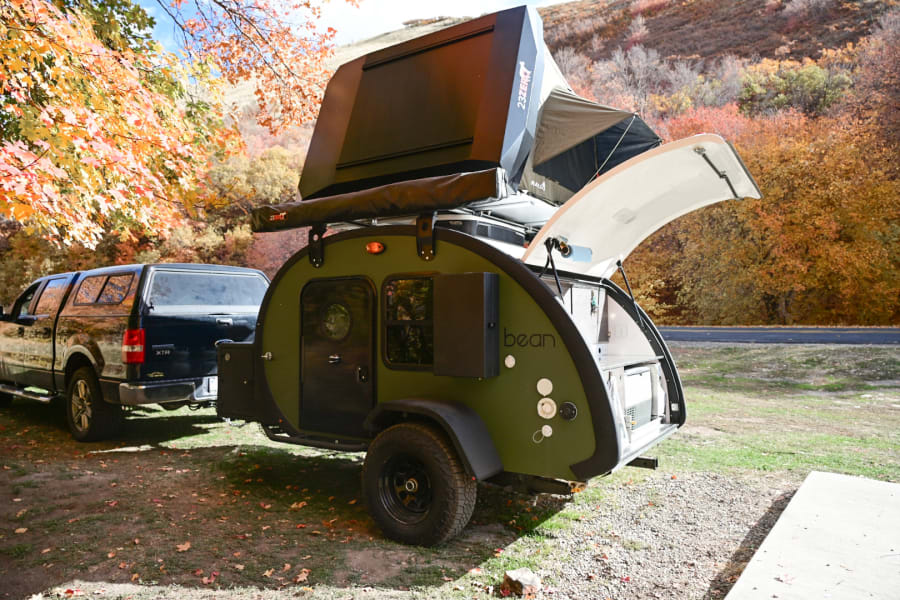 A green Bean teardrop trailer with rooftop tent and truck. Ideal RV rental for fall camping amidst vibrant autumn leaves.