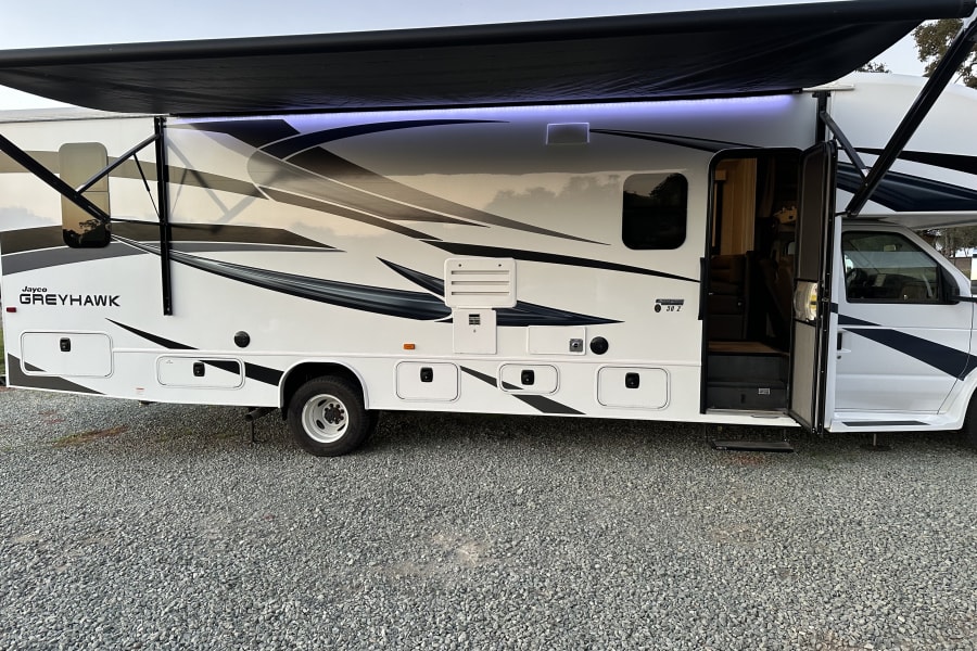 White Jayco Greyhawk RV rental with an extended black awning, purple LED lights, and an open entry door parked on gravel.