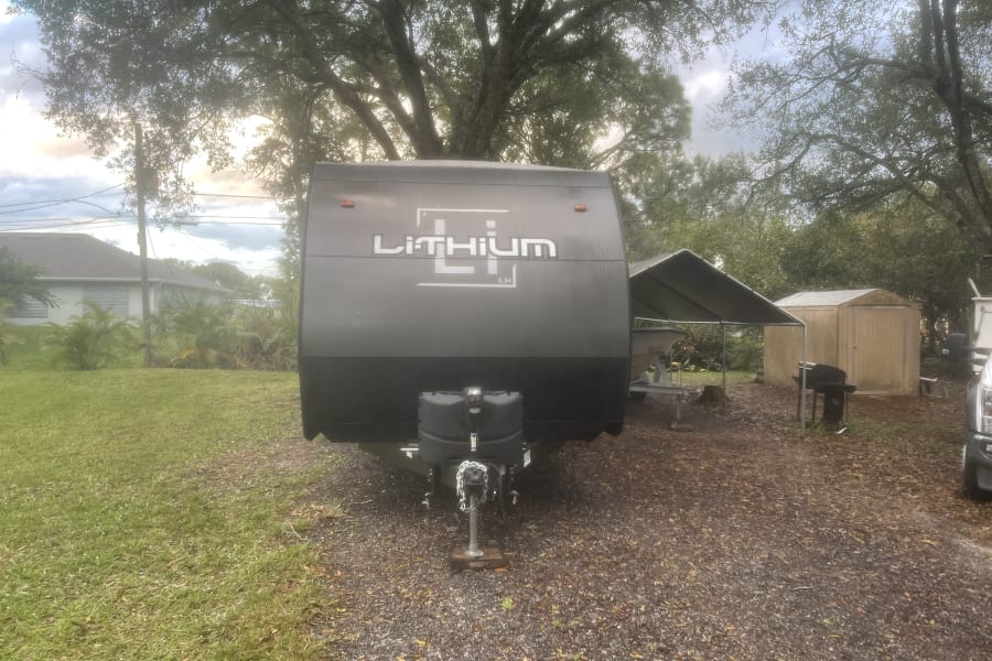 Dark gray LiTHIUM RV rental on gravel, surrounded by trees and grass, with a house and shed in the background.