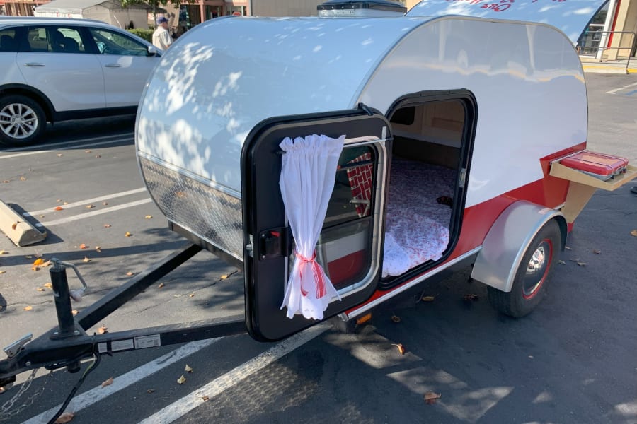 Cozy white & red teardrop camper, an ideal RV rental, with an open door showing the bed & small pull-out stove for travel.