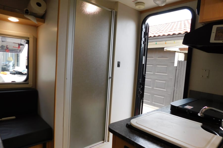 Interior view of an RV rental showing the kitchen with a sink, cutting board, a frosted shower door, and an open entry door.
