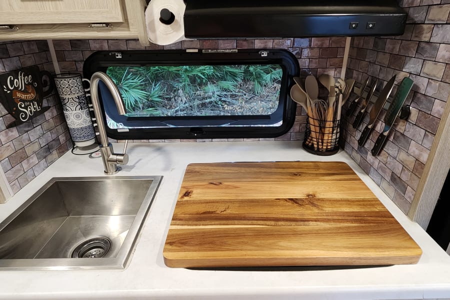 This RV rental kitchen features a bright counter, stainless steel sink, wood cutting board, utensil storage, and a scenic window view.