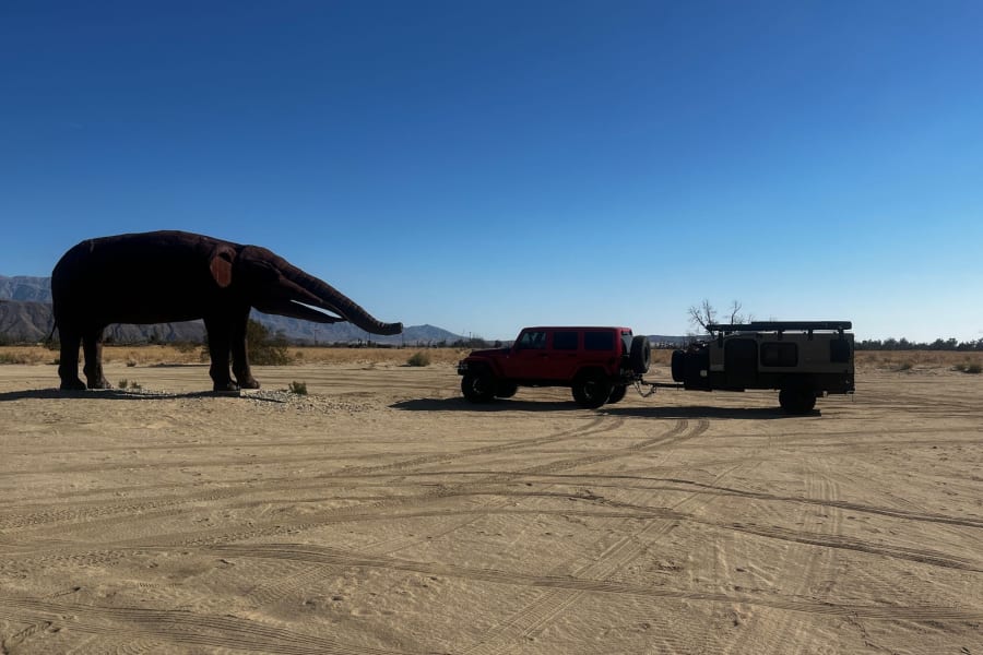 A red Jeep pulls an off-road RV rental trailer, parked near a giant elephant sculpture in the desert under a clear blue sky.