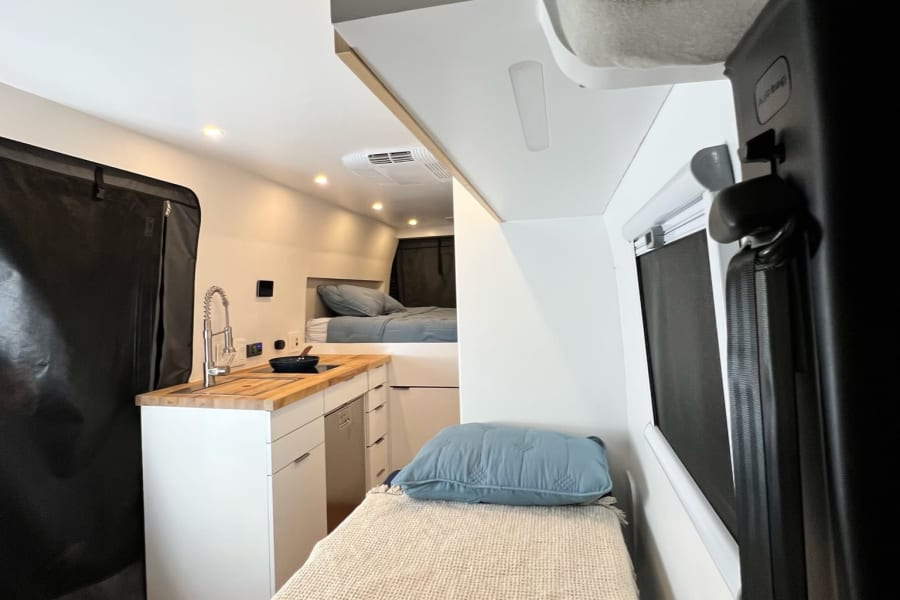 Bright RV rental interior: compact kitchen with wood counter/sink, plus two cozy bed areas visible.