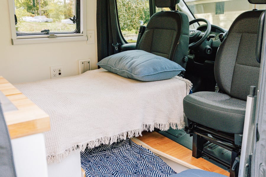 Inside a modern rv rental: a cozy bed, pull-out floor cushion, light wood cabinets, and clear view to the front cabin.