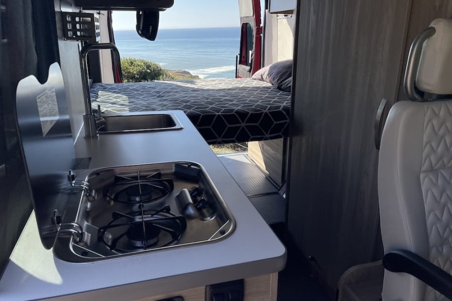 Inside an RV rental: Compact kitchen, comfy bed, and a stunning ocean view perfect for your next road trip.