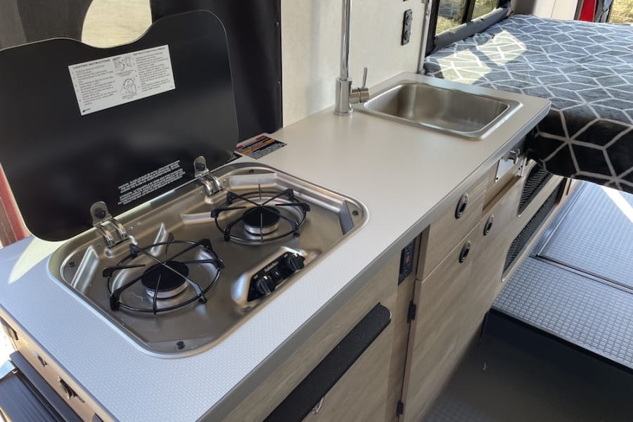 Inside an RV rental: Compact kitchen with a two-burner stove, sink, and counter, next to a bed with a geometric blanket.