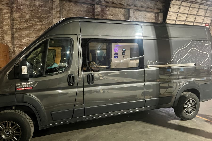 Gray NOKE Van Co. Ram ProMaster camper van with topo graphics, equipped for RV rental journeys. See inside controls.