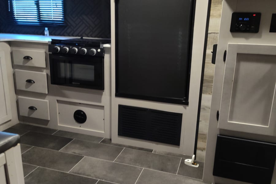Modern RV rental interior: kitchen with microwave, fridge, oven, light cabinets, and dark tile floor.