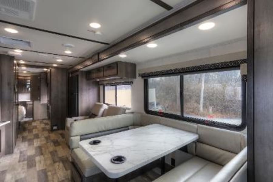 Spacious RV rental interior featuring a dinette with bench seating, a sofa, and windows showing outdoor scenery.