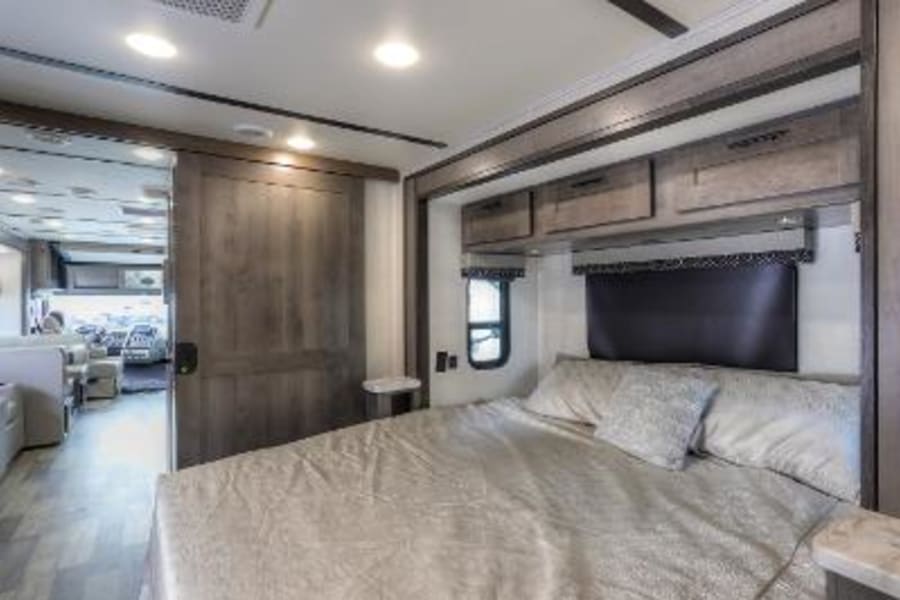Comfortable RV rental bedroom featuring a queen bed, overhead storage, and a sliding door connecting to the living space.