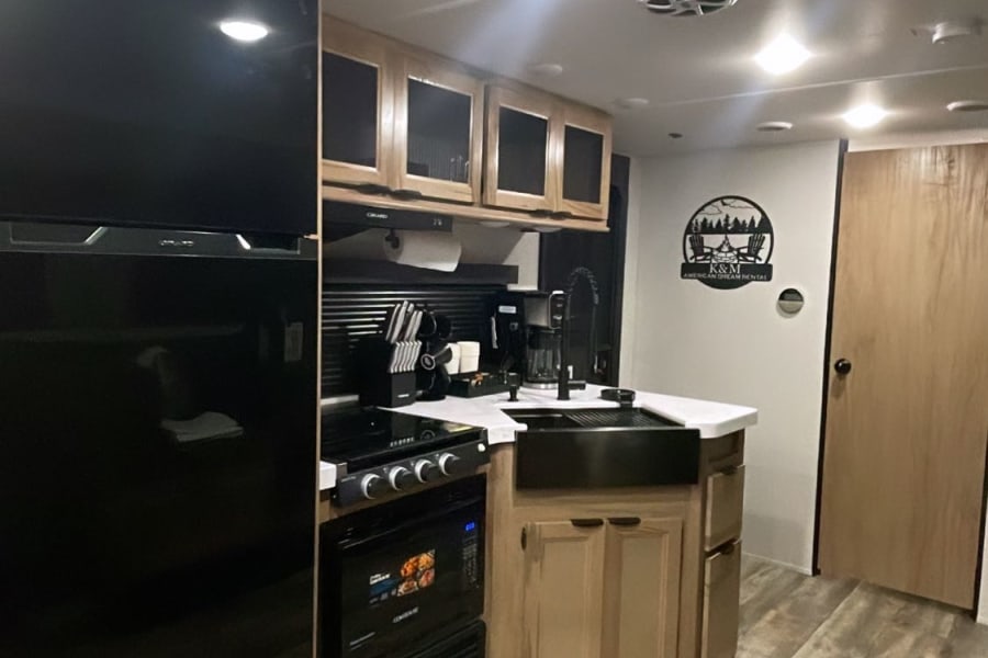 Modern RV rental kitchen featuring black appliances, wood cabinets, sleek flooring, and a cozy decor sign.