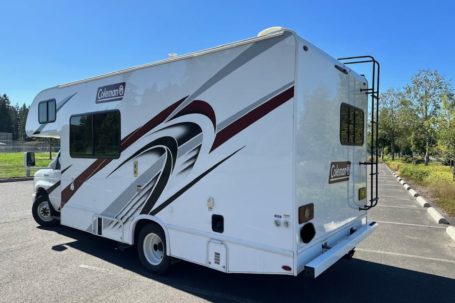 Explore the outdoors with this white Coleman RV rental. This motorhome features graphics and a rear ladder, parked on asphalt.