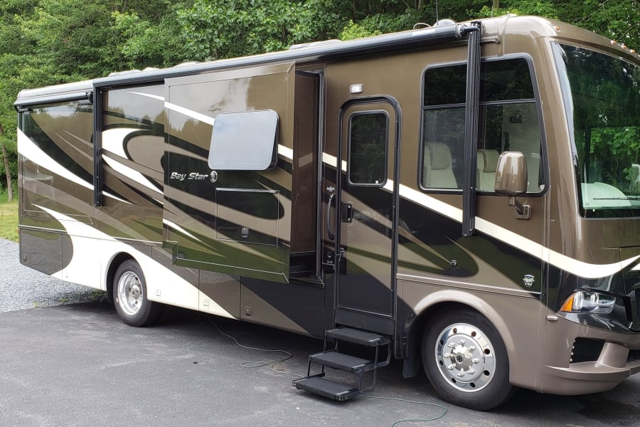 A large brown and white Newmar Bay Star motorhome for rv rental, parked on asphalt with a lush green forest background.