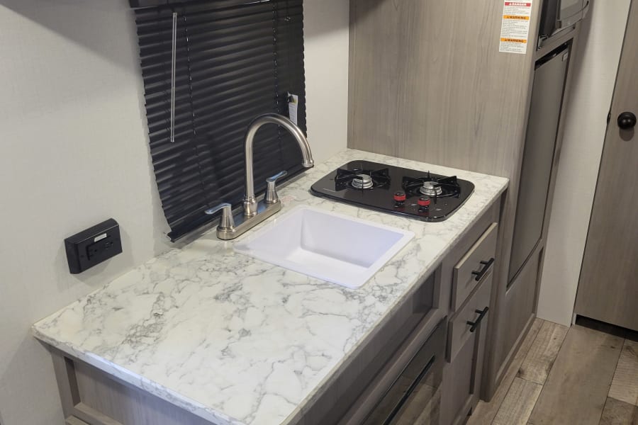 Compact RV rental kitchen features sink, stovetop, microwave, and cabinets. Ideal travel trailer interior.