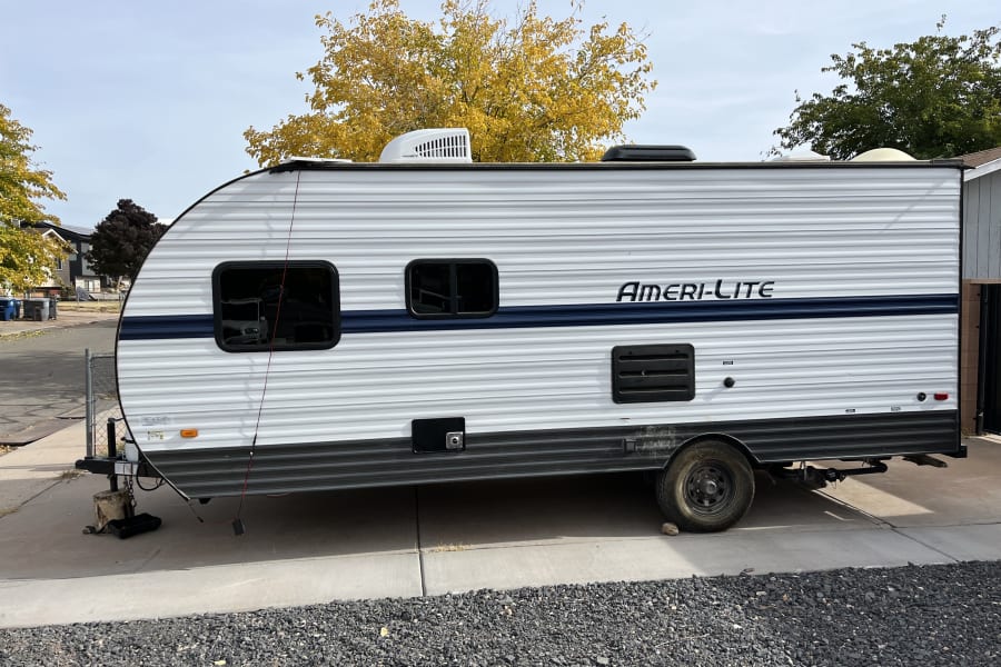White Ameri-Lite travel trailer RV rental, with a blue stripe, parked on concrete next to gravel and fall trees.