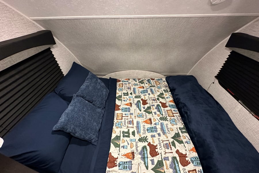 Cozy RV rental bedroom with dark blue bedding and a nature-themed quilt, featuring compact design and window blinds.