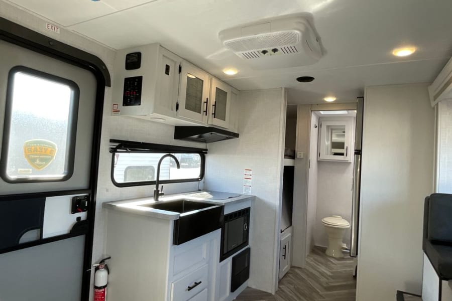 Explore this rv rental's interior: a modern kitchen area with sink, microwave, and fridge, leading to a bathroom and bunks.