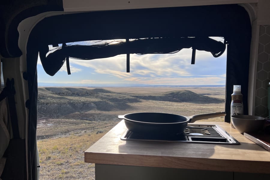 Inside an RV rental, a pan sits on a stovetop with olive oil nearby. The large window reveals a vast, hilly desert landscape.