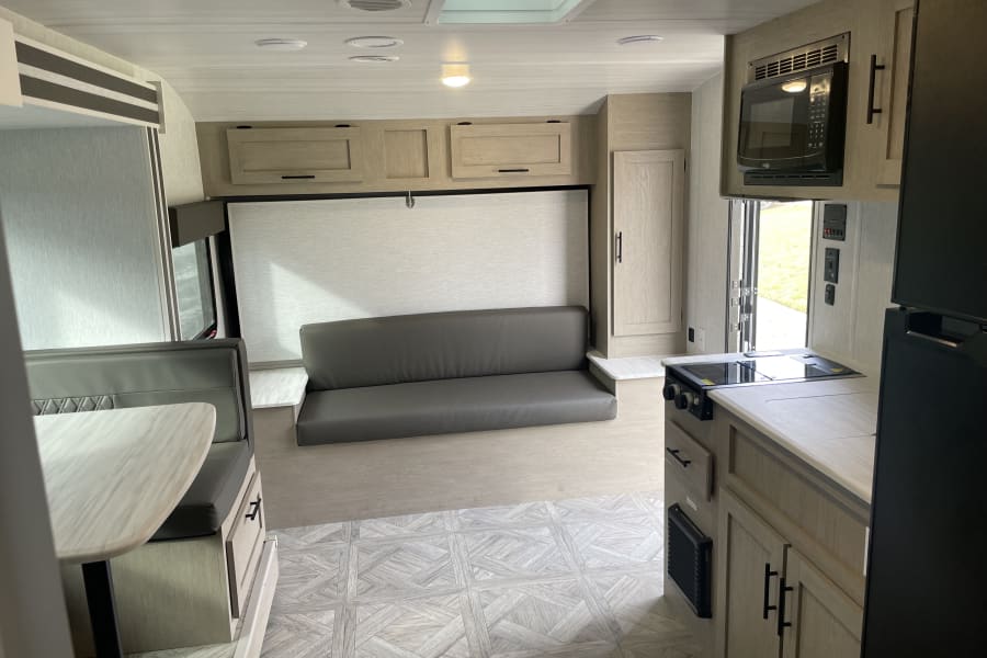 Inside a modern RV rental, featuring a full kitchen with microwave, dinette, and a sofa. Light wood cabinetry and patterned floor.