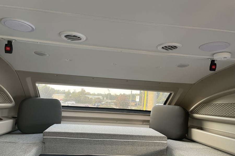 Inside an RV rental: a spacious overhead sleeping area with light grey cushions, ceiling vents, and window view.