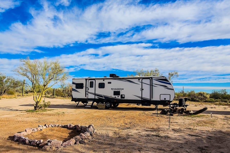 This spacious **RV rental** offers desert camping with a fire pit, parked on sandy ground under a vibrant blue sky.