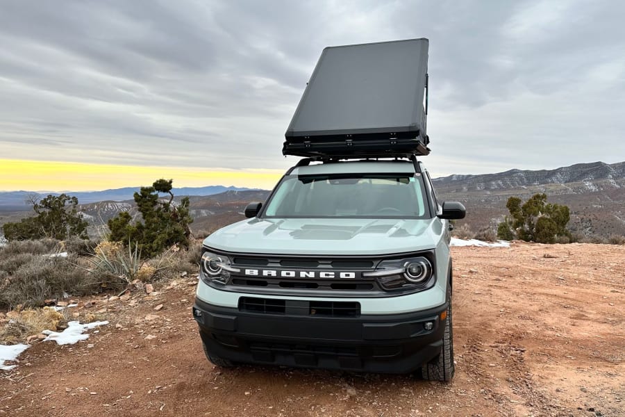 Stunning view! A light gray Ford Bronco Sport with a deployed rooftop tent, parked off-road overlooking mountains. Consider this versatile rv rental.