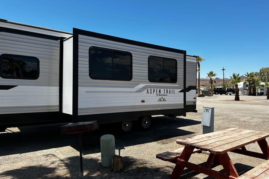 Discover your next adventure! This RV rental, an Aspen Trail travel trailer, is set up at a sunny campsite with a picnic table.