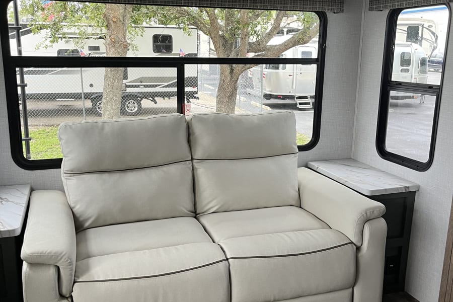 This RV rental interior shows a cream two-seater sofa, marble-top side tables, and dark cabinets. Windows offer an RV lot view.